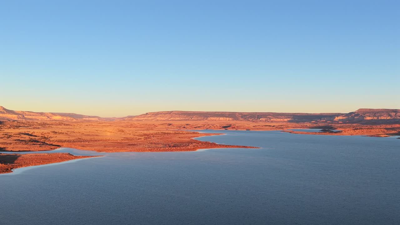 Breathtaking aerial footage captures the sunrise over Abiquiu Lake, New Mexico, bathing the rugged red rock and high desert landscape in a warm, golden light. Perfect Southwest morning scenery