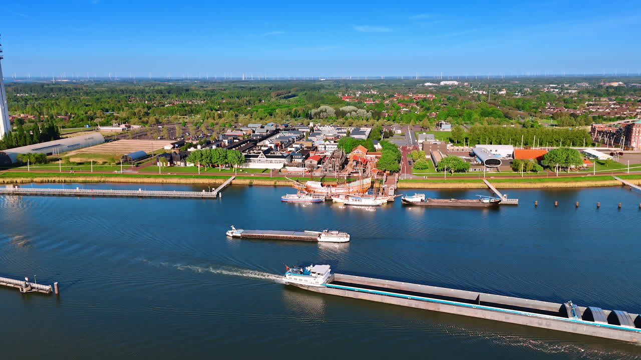 Unloaded barges float by the calm waterscape along the dams. Aerial view on the lakefront of Lelystad, the Netherlands, Batavialand museum and telecom tower.