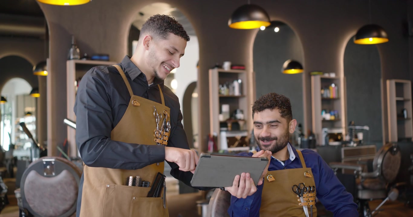 Barbieri uomini afroamericani con grembiuli color cachi in salone che toccano un tablet, condividendo idee per tagli e ridendo.
