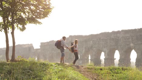 joven pareja encantadora mochileros turistas caminando de la mano llegan a la colina frente al acueducto romano arcos en el parco degli aquedotti ruinas del parque en roma en romántico amanecer brumoso con guitarra y saco de dormir cámara lenta