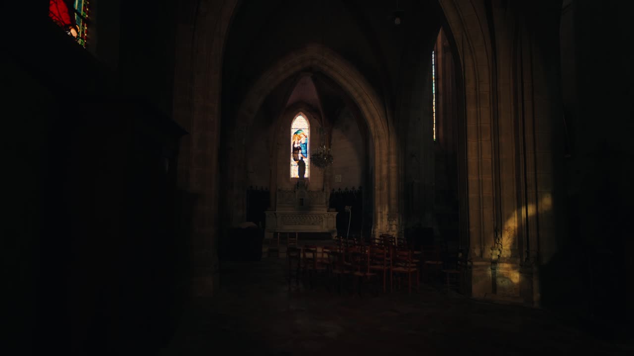 A luz do sol filtra-se através de um vitral para o interior escuro e os arcos de pedra de uma igreja histórica em Issigeac, Dordogne. Vista em ângulo baixo