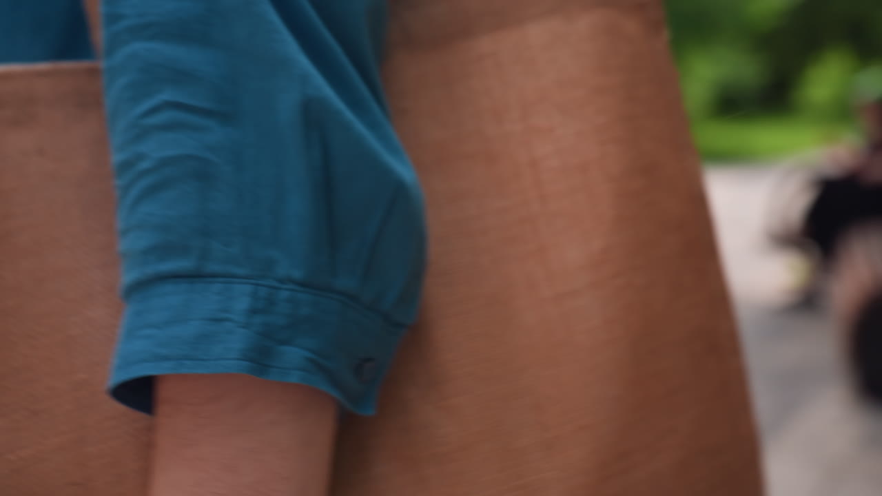 Close up of woman in blue shirt lifting brown tote bag from wooden bench in sunny park, preparing to walk away, soft natural light enhancing texture of fabric and wood