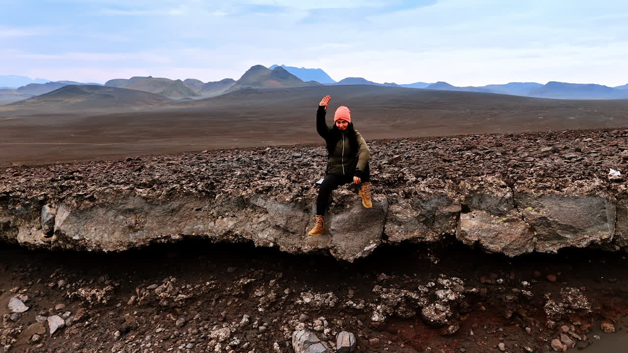 Woman sits on the rock edge waving her hand. Drone distances from tourist resting among the cosmic scenery of Iceland.