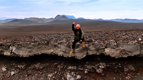 woman sits on the rock edge waving her hand. drone distances from tourist resting among the cosmic scenery of iceland.
