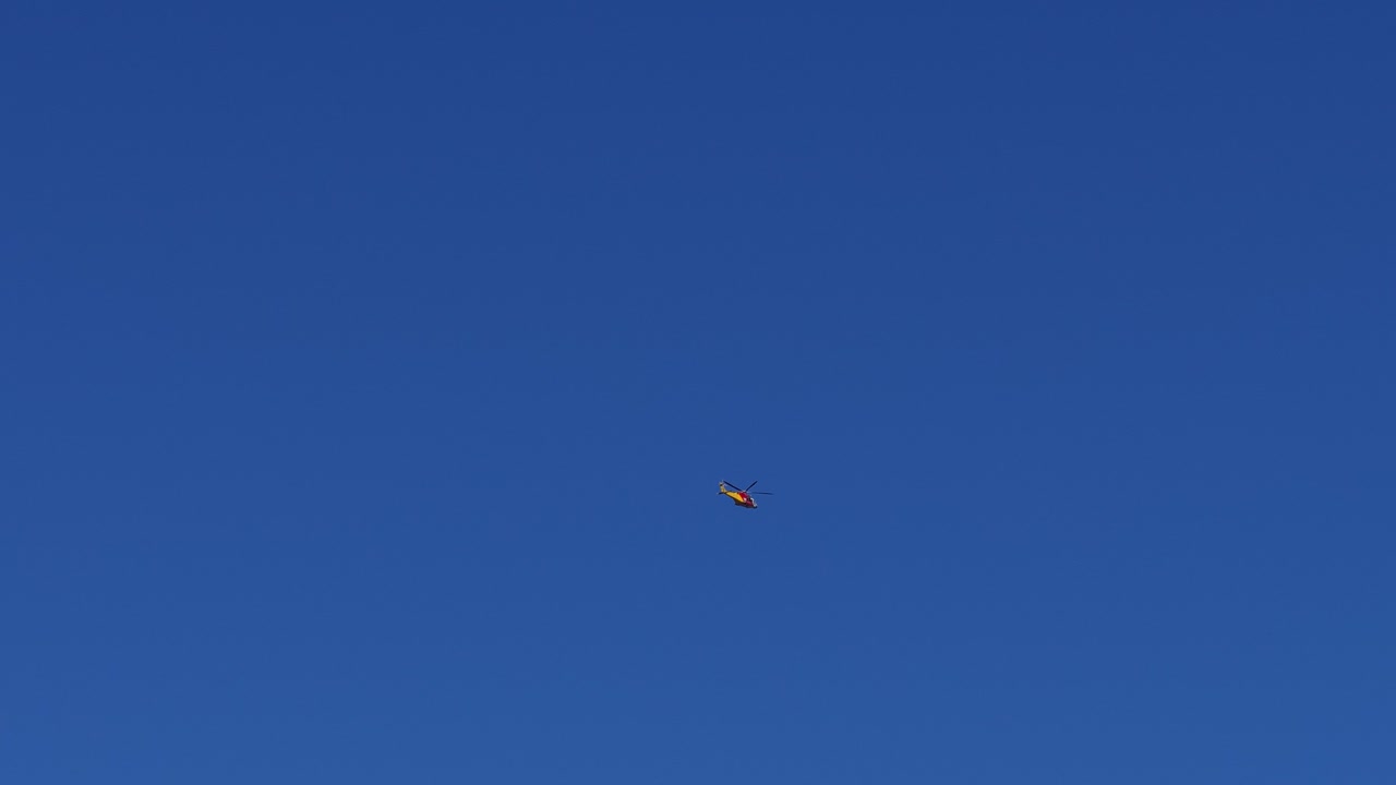 A rescue helicopter steadily flies through a cloudless blue sky, captured in wide static shots