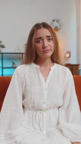 portrait of upset young woman in white dress sitting on sofa showing heartbroken sign with her hands