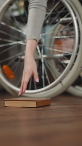 mujer mueve la silla de ruedas tratando de alcanzar un libro tendido en el suelo. persona femenina con discapacidad experimenta dificultades para recoger objetos de cerca