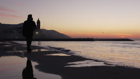 man walks along tranquil sea, dusky coastline stretches out, leading towards silhouette of a church set against the majestic mountain range