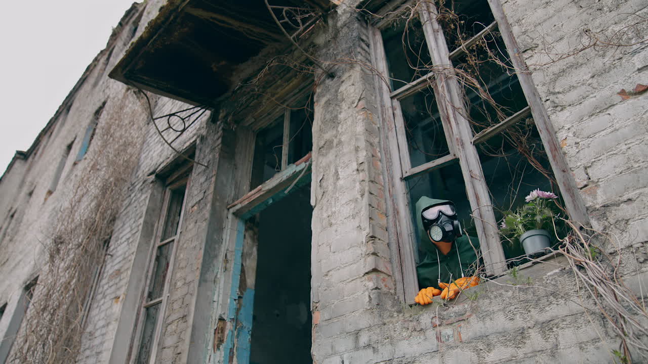 Human in respirator looking from ruined building with broken windows. Flower pot and ecologist in the abandoned place after biological attack.