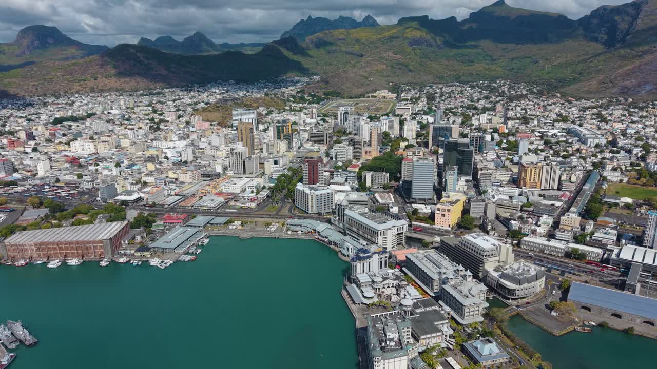 Pull out drone shot of Port Louis skyline in Mauritius. Features the bustling Caudan Waterfront, with the city nestled against the Moka Mountains and the blue ocean
