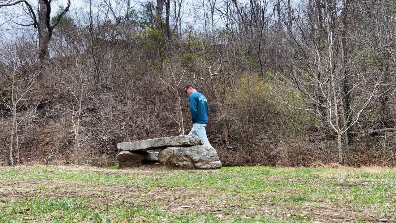 파란색 옷을 입은 젊은 백인 남성이 삶을 생각하기 위해 공원 한가운데 있는 조용한 바위 벤치로 천천히 걸어갑니다.