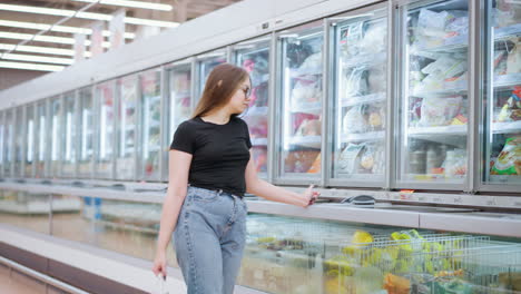 young woman shopping in mall, stops at fridge display, places hand on glass then continues walking with shopping bags in a brightly lit store, modern retail environment with frozen food section