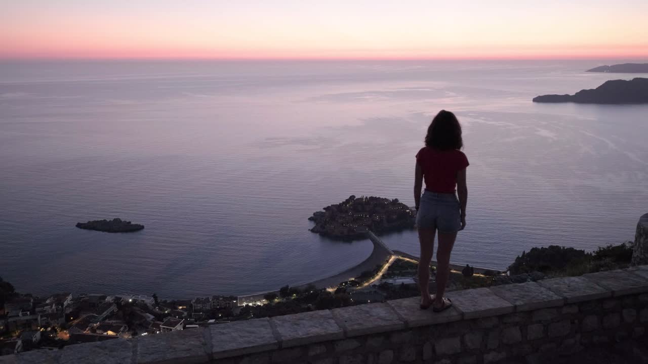 Beautiful lady walking on a viewpoint looking over the Montenegro coastline over top of Sveti Stefan during a beautiful Sunset