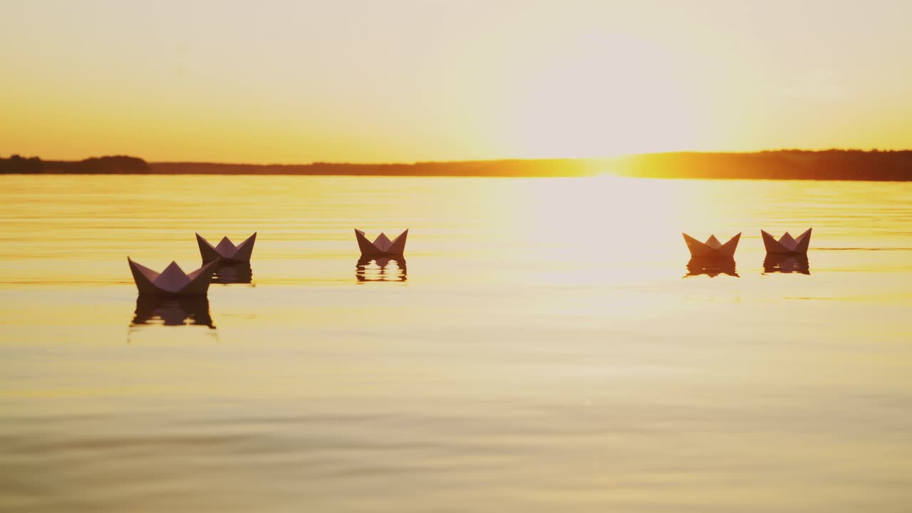 Yellow rays of the evening sun shining on the paper boats swimming in the river at sunset. Origami ships reflects in the water outdoors