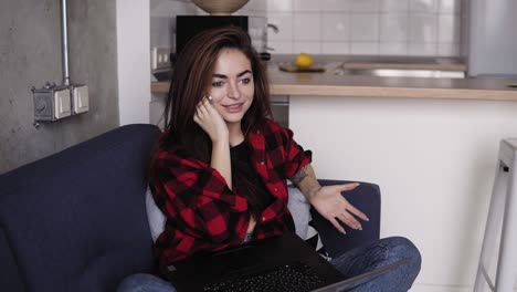 Una-Chica-Increíblemente-Hermosa-Hablando-Con-Alguien-Por-Teléfono-Mientras-Sostiene-Una-Computadora-Portátil-De-Rodillas,-Sentada-En-Un-Sofá.