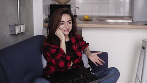 una chica increíblemente hermosa hablando con alguien por teléfono mientras sostiene una computadora portátil de rodillas, sentada en un sofá.