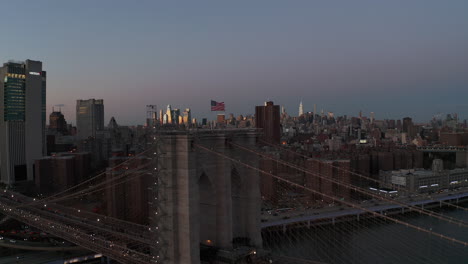 Vorwärts-Fliegen-Um-Die-Brooklyn-Bridge-Mit-Der-Amerikanischen-Flagge-An-Der-Spitze.-Wohnbezirk-Mit-Mehrfamilienhaus-In-Der-Abenddämmerung.-Beleuchtete-Wolkenkratzer-In-Der-Ferne.-Manhattan,-New-York-City,-Vereinigte-Staaten