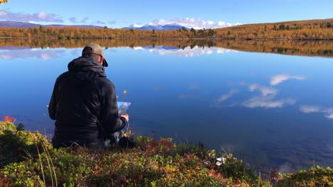 Hiker-reading-his-map-sitting-by-the-edge-of-a-crisp-clear-mountain-lake-during-autumn-in-Abisko,-Lapland,-Sweden