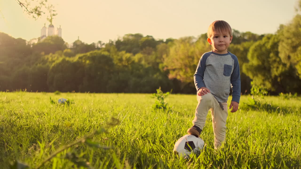 un niño de pie en la hierba con una pelota de fútbol al atardecer.