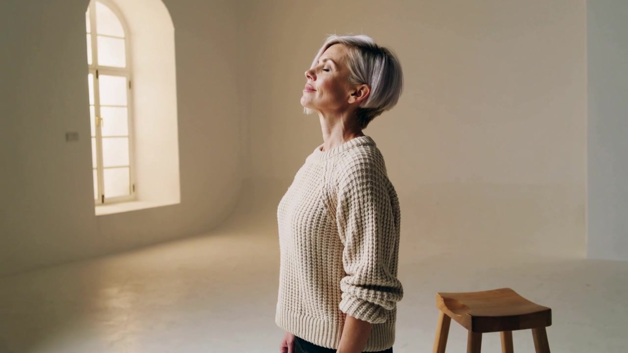 Donna anziana che respira, sollevando il mento, mantenendo una posa calma in uno studio essenziale, con maglione di lana, sgabello e finestra.