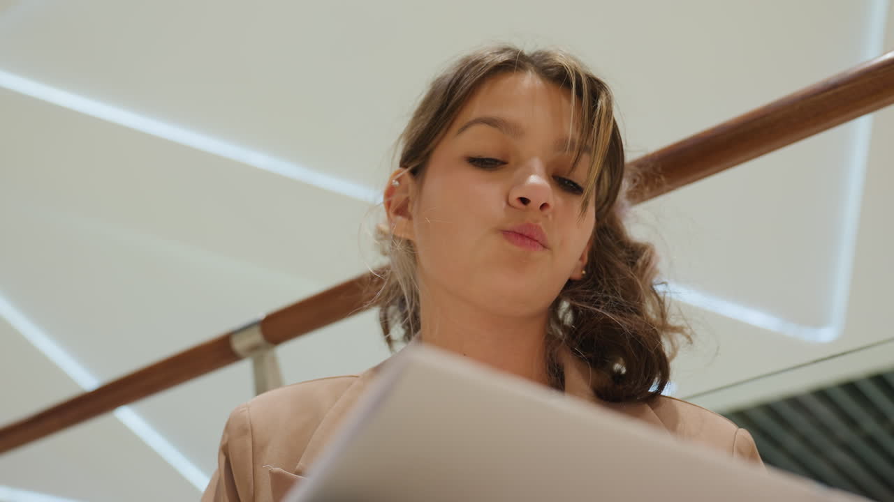 쇼핑몰에서 노트북을 들고 올려다보며, 부드러운 조명과 현대적인 난간이 있는 곳에서 공상에 잠겨 계획을 세우는 여성, 창의력과 공공장소가 어우러져 영감을 주는 스토리텔링을 위한 반사적인 분위기