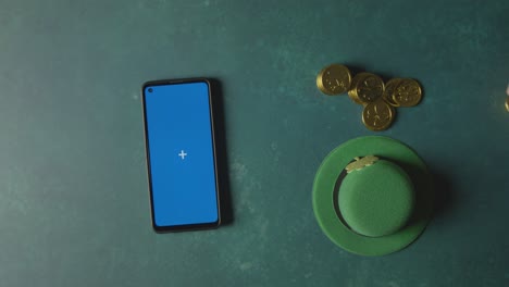 overhead studio shot of green leprechaun top hat with gold coins and blue screen mobile phone to celebrate st patricks day 1