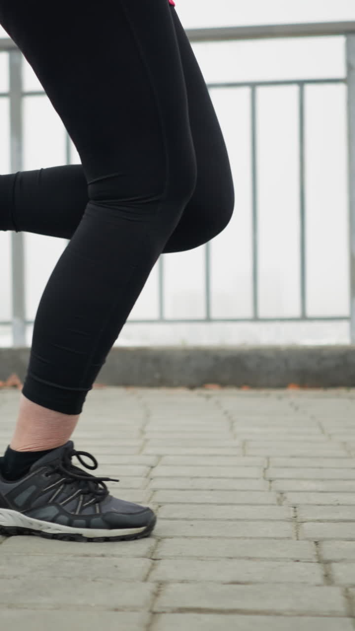 leg view of lady in black sneakers and leggings jogging near iron railing of bridge overlooking foggy city view on snowy pathway during winter outdoor fitness movement showcasing endurance