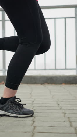 leg view of lady in black sneakers and leggings jogging near iron railing of bridge overlooking foggy city view on snowy pathway during winter outdoor fitness movement showcasing endurance