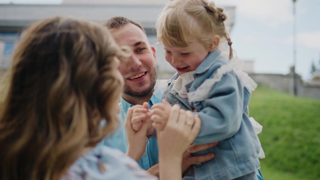 Junge Eltern interagieren freudig mit ihrer kleinen Tochter im Freien. Der Vater hält das Kind und lächelt warmherzig, während sie kichert und spielt. Die Mutter gesellt sich dazu und berührt die Hand des Kindes