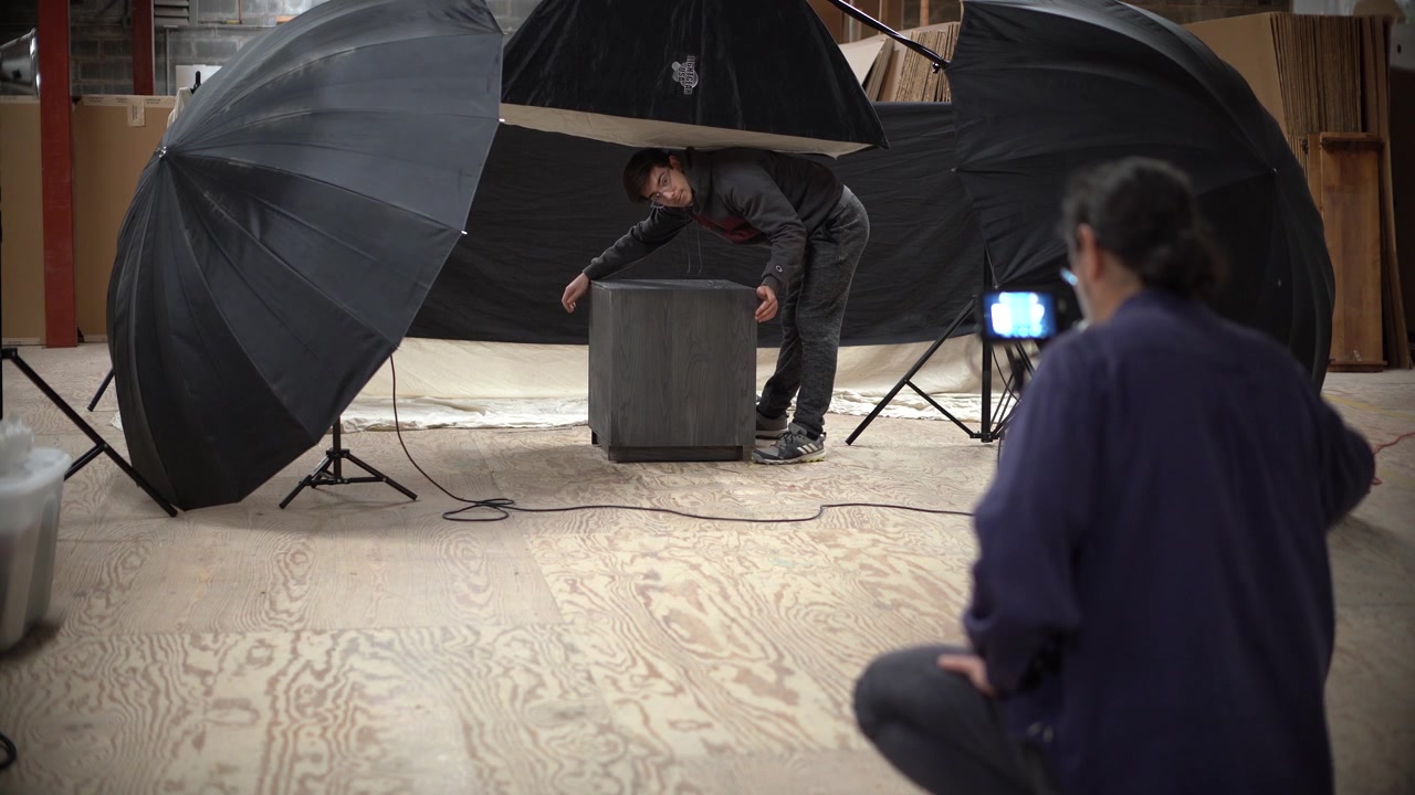 Muž fotografuje noční stolek na fotografickém place a asistent otáčí nábytkem pro jiný úhel
