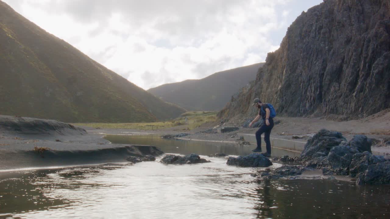 un excursionista de un día en solitario caminando cuidadosamente de roca en roca a través de un río costero con un valle soleado en el fondo