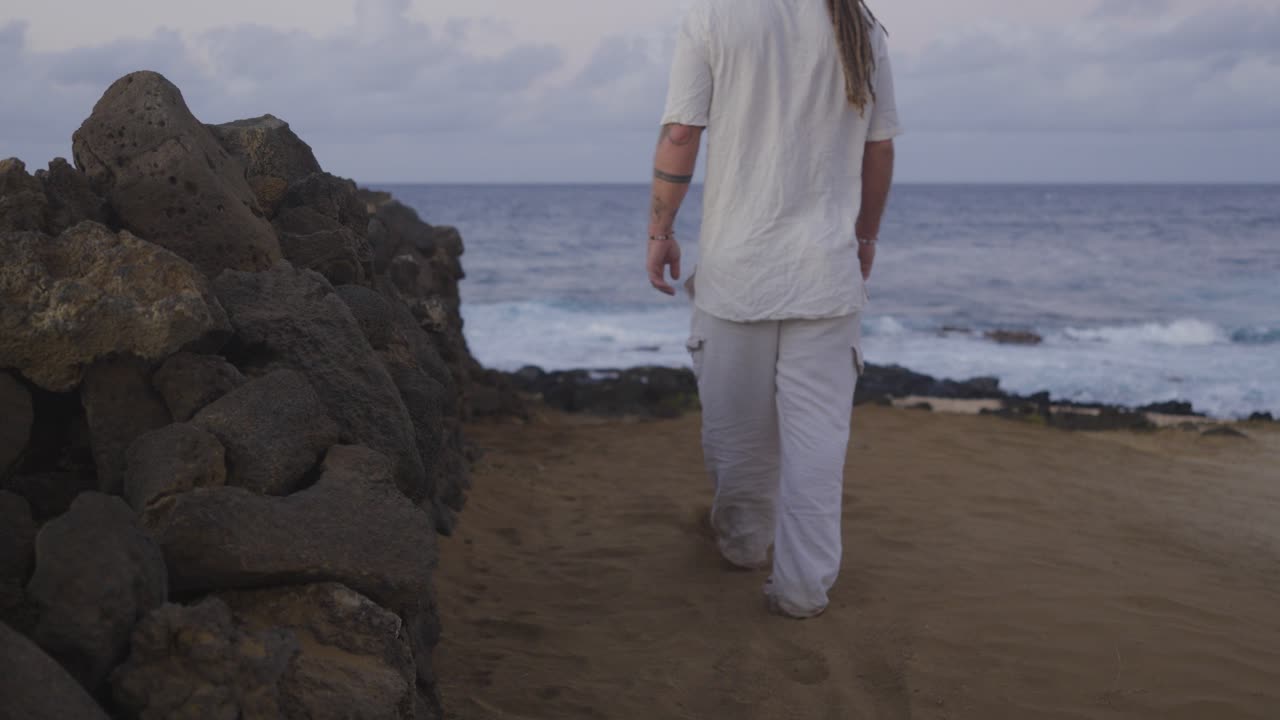 Dressed in loose white clothing, a man walks away from the camera along a rugged sandy path bordered by a stone wall, with the ocean waves and horizon ahead of him