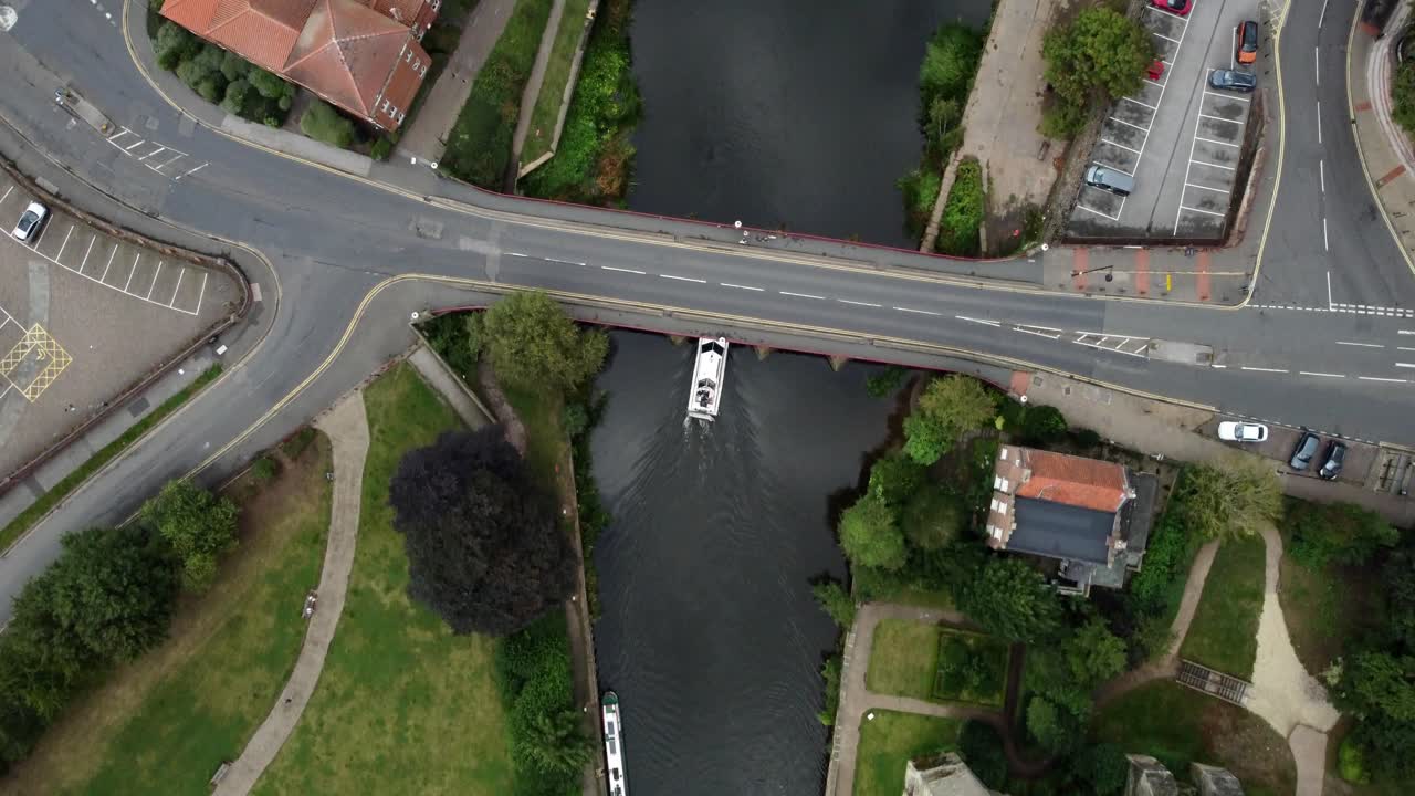 Drone video follows directly above looking down on a River Boat heading along the River Trent in Newark. This captures the boat head under a road bridge with cars driving on and carparks around.