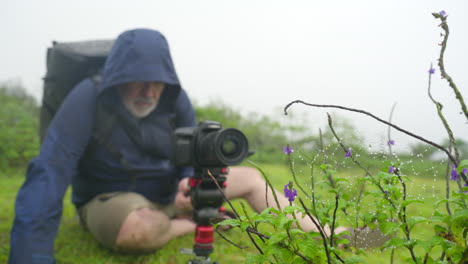 viejo fotógrafo de naturaleza en chaqueta con capucha establece una cámara montada en un campo de hierba