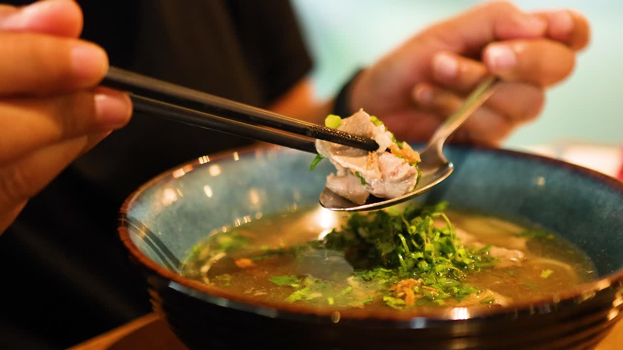 A person skillfully uses chopsticks and a spoon to enjoy a bowl of Vietnamese pho in a restaurant setting