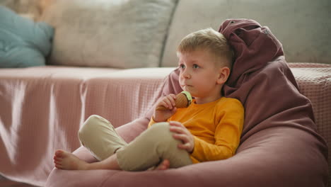 young boy sitting barefoot on pink bean bag chair indoors happily eating mint chocolate ice cream cone, wearing yellow long-sleeve shirt, looking ahead