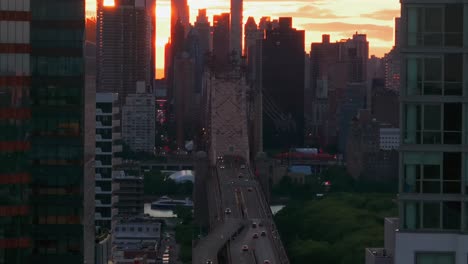 an aerial shot of the queensboro bridge, seen from long island city between skyscrapers in queens during a golden sunset. the drone camera dolly out and tilt up slowly revealing more buildings.