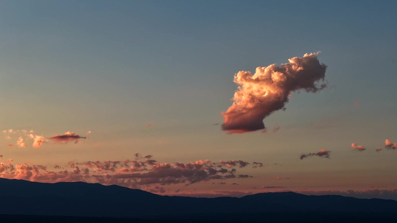 4K drone timelapse of a single, picturesque cumulus cloud illuminated by stunning pink and orange sunset colors. The isolated cloud drifts over a dark, silhouetted high desert mountain range.