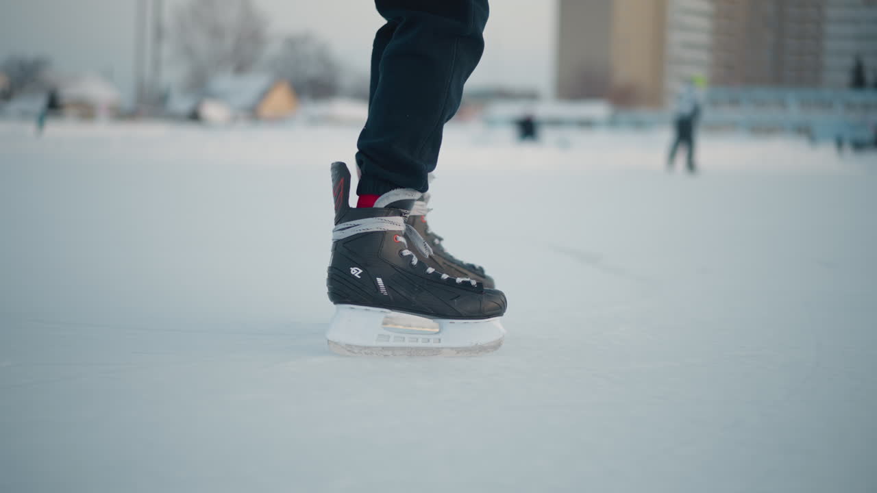 leg view of person skating past another on frozen ice rink with blurred people and building in background under soft winter light, ice surface textured with skate marks and snow around edges