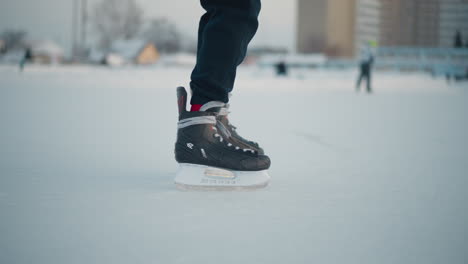 leg view of person skating past another on frozen ice rink with blurred people and building in background under soft winter light, ice surface textured with skate marks and snow around edges