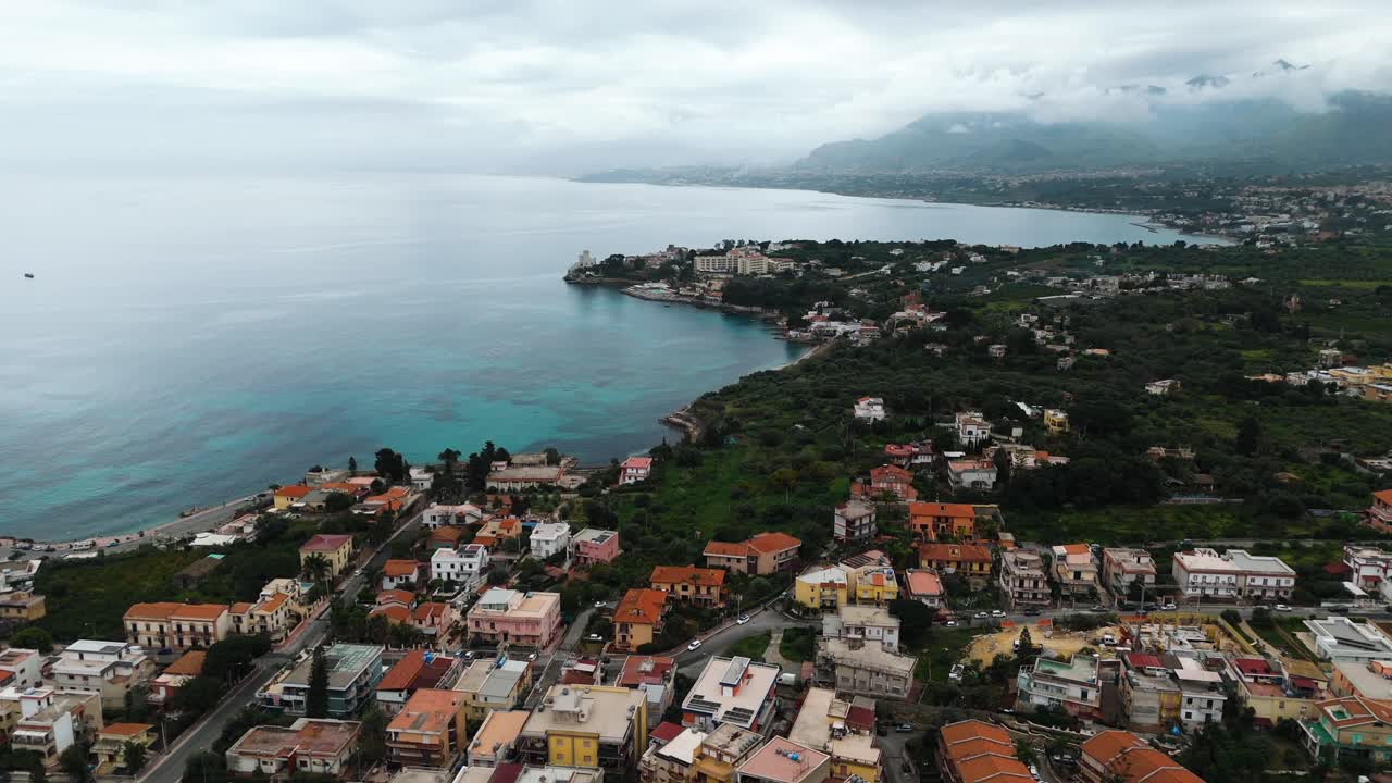 Drone flight along the northern Sicilian coast near Casteldaccia and Santa Flavia showing residential buildings, green hills, and coastline with turquoise water and cloud-covered mountains