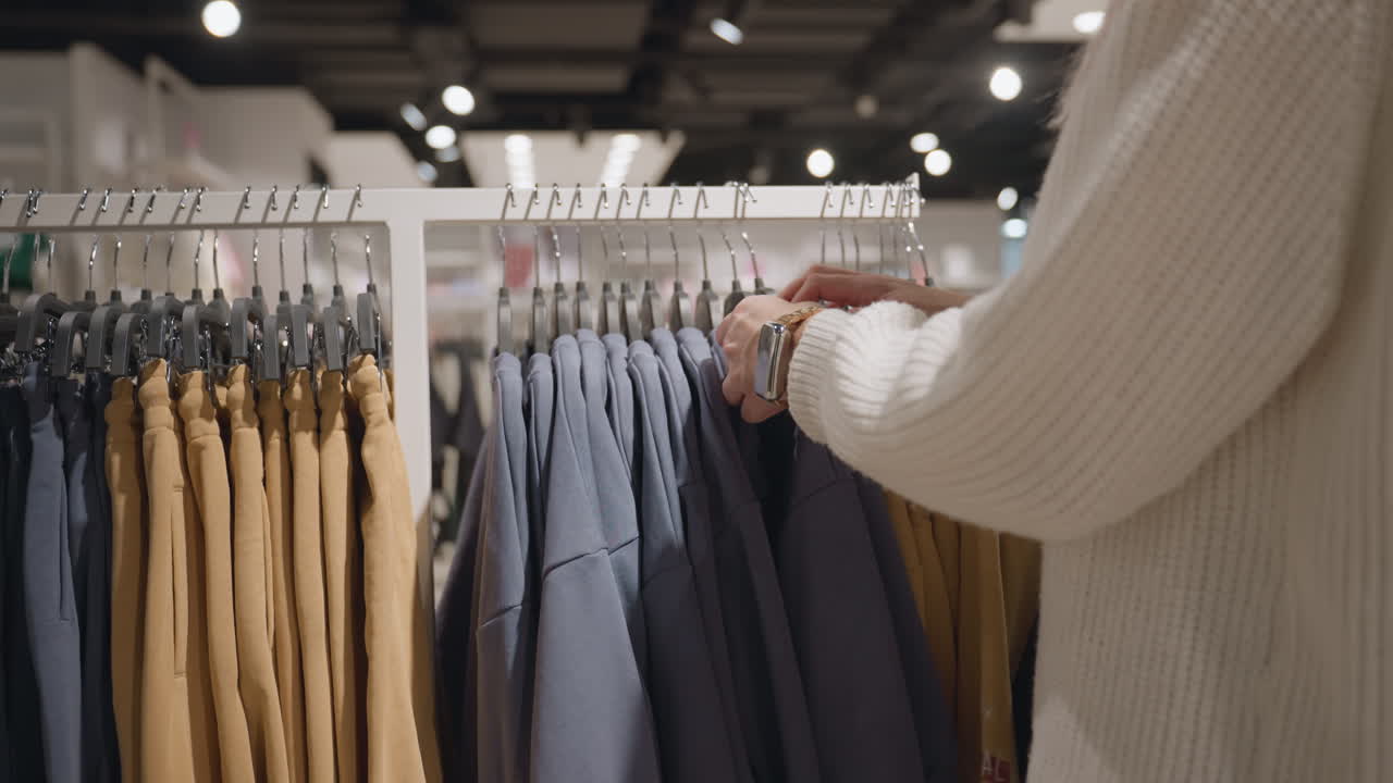Well lighted boutique showcases neat row of camel sweatpants and slate blue polos on metal rack, hangers glint under ceiling lamps while curious shopper hand with lavender manicure hovers to browse