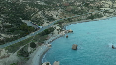 aerial view of a coastal road meandering along a rugged shoreline dotted with boulders, next to a clear turquoise sea, cars parked along a small beach inlet and hilly terrain in the background pafos