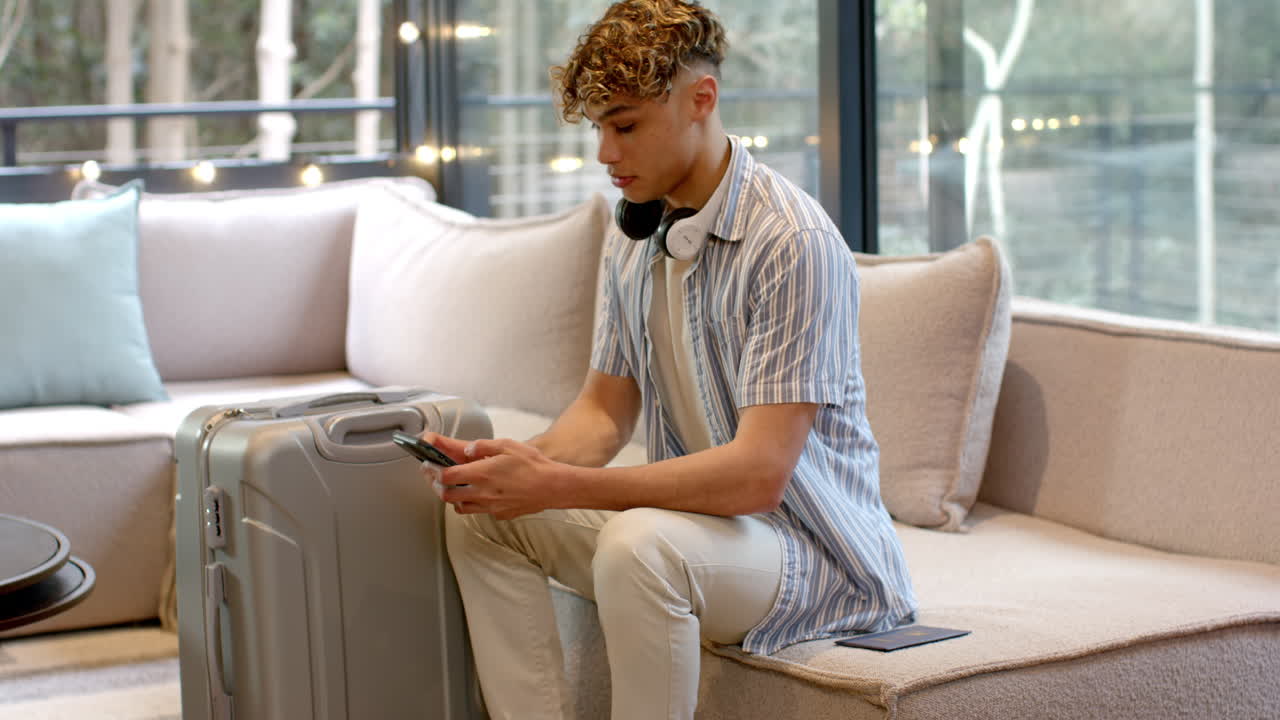 Young man with headphones using smartphone, sitting on sofa with suitcase
