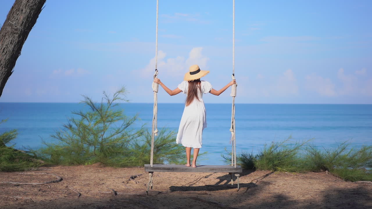 모자를 쓰고 바다를 바라보며 바닷가 그네를 타고 있는 젊은 여성의 뒷모습