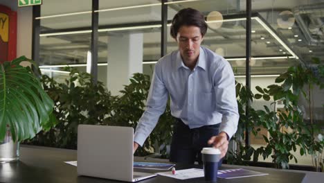 Caucasian-businessman-standing-using-laptop-going-through-paperwork-in-modern-office