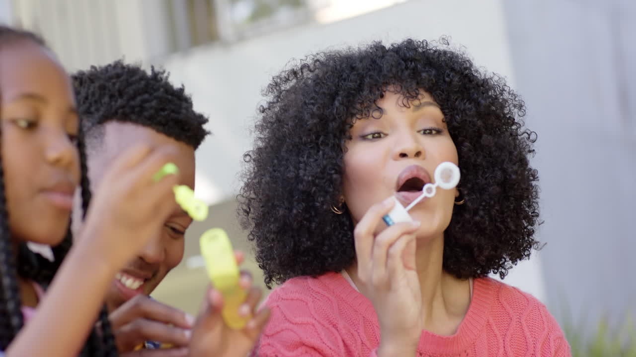 Happy african american parents with daughter blowing bubbles in garden at home, slow motion