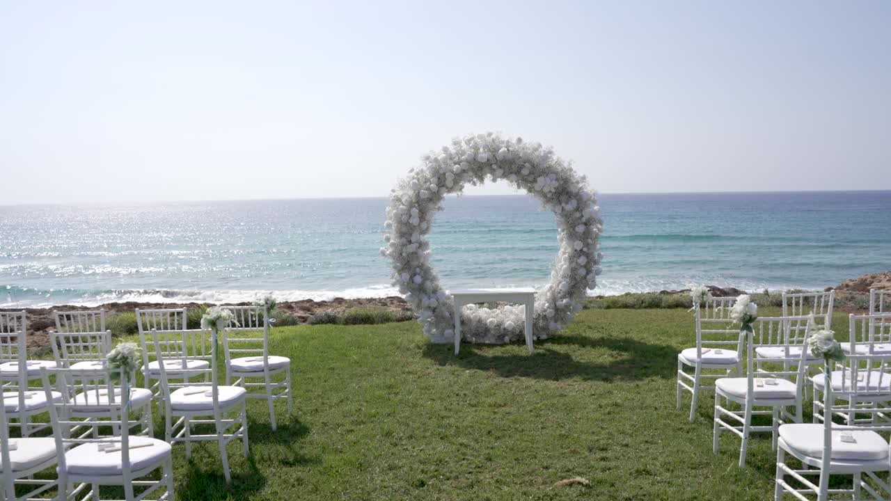 Ci sono delle sedie disposte sull'erba vicino all'oceano, con un arco floreale posizionato di fronte. Il sole splende e le onde si infrangono dolcemente. Questa è una giornata luminosa per un evento speciale.