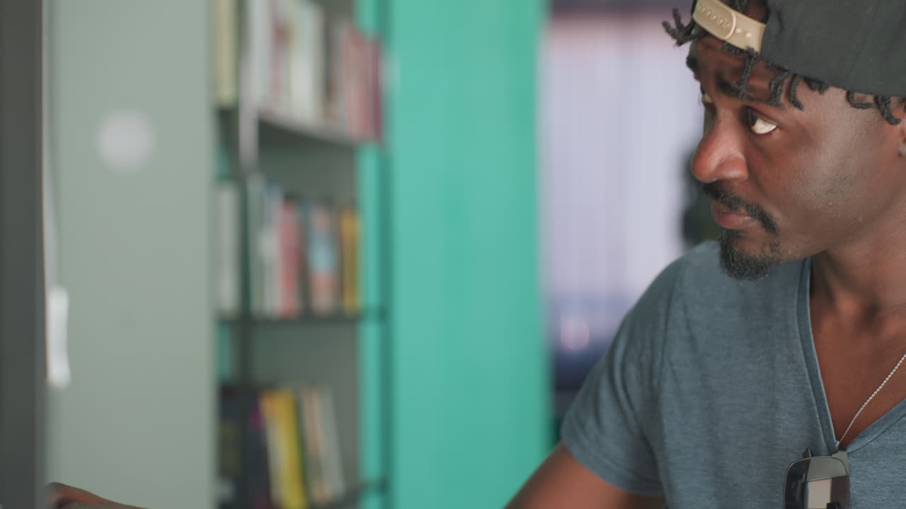 Joven africano vestido con ropa informal, gorra hacia atrás y gafas de sol en la camisa, de pie en el pasillo de una biblioteca, sosteniendo un libro abierto y leyendo atentamente junto a estanterías llenas de libros
