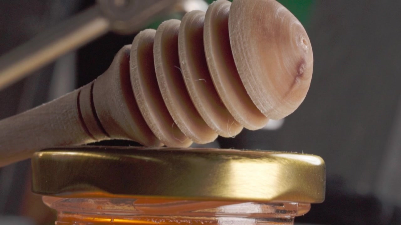Macro primo piano che mostra un mestolo di legno per miele appoggiato sopra un barattolo di vetro aperto pieno di miele dorato, evidenziando la consistenza lucida, il colore caldo e i dettagli fini
